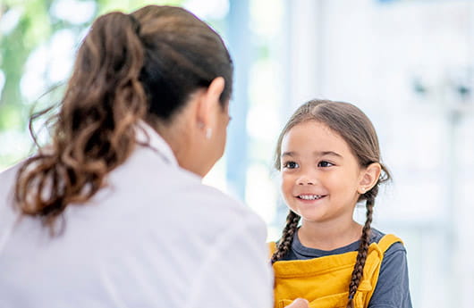 A cute little girl with pigtails smiles while a healthcare professional talks to her.