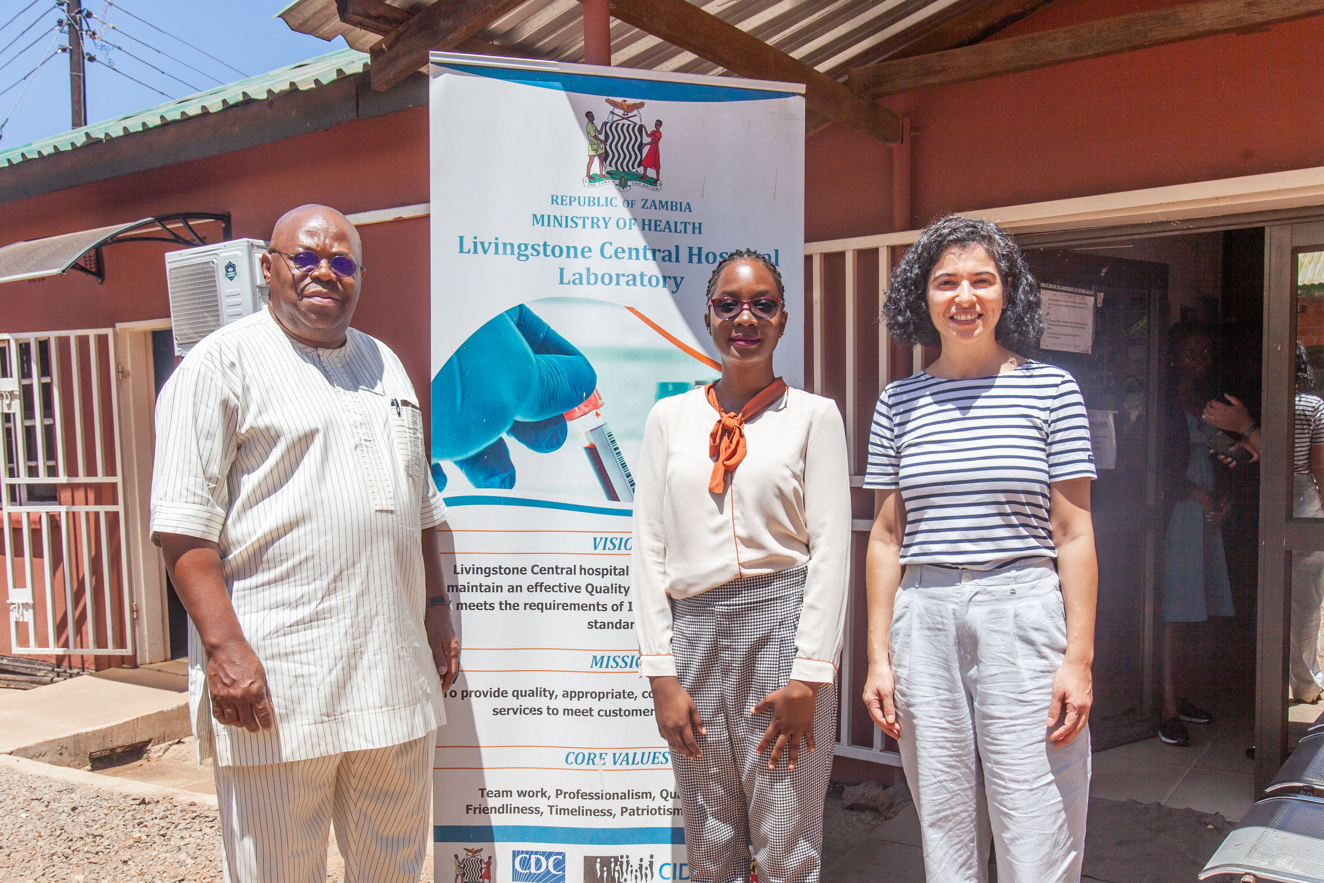 Three laboratory medicine professionals stand outside the entrance to a workshop in Zambia.