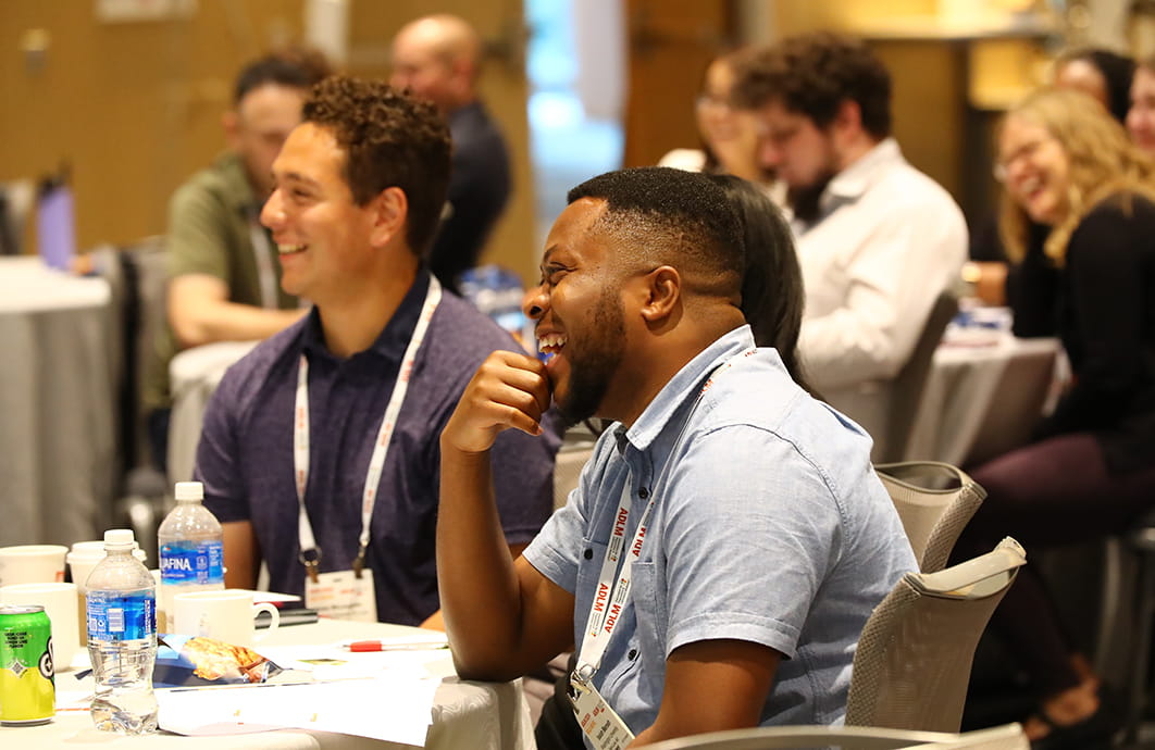 Two men sitting at a table and smiling while watching a presentation.